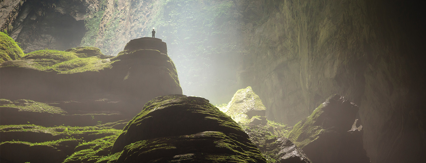 A person stands atop a moss-covered rock formation inside a vast cave with light filtering from above